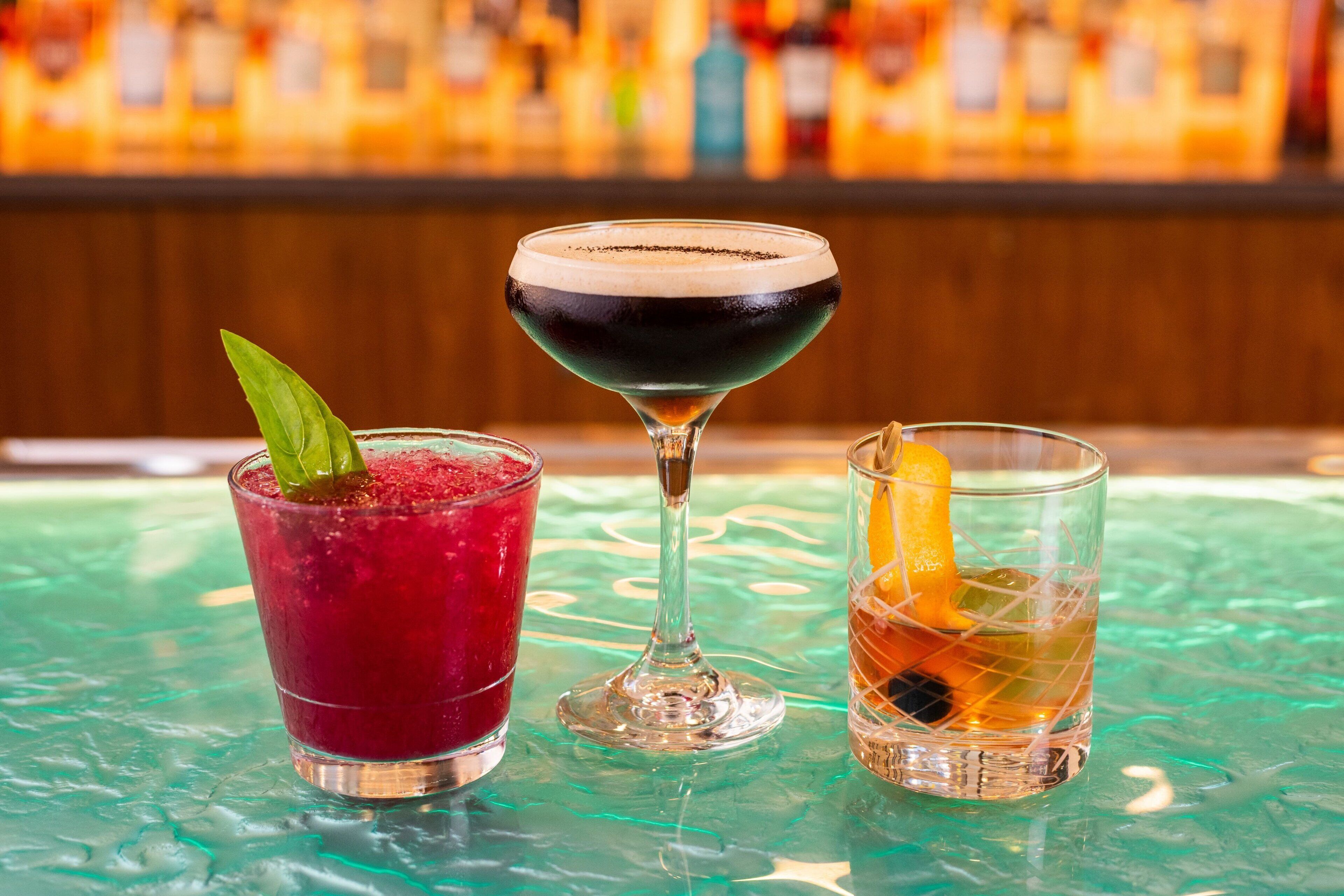Three drinks on a bar counter with a blurred background of bottles.