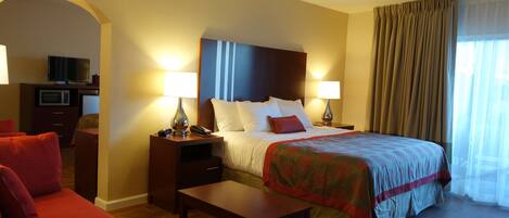 Habitación de hotel con cama, dos mesitas de noche con lámparas, televisor, microondas, sofá rojo y suelo de madera.