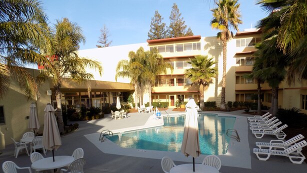 A pool area with lounge chairs and tables, surrounded by palm trees and a building with multiple balconies. - Ramada by Wyndham Sacramento (Sacramento)