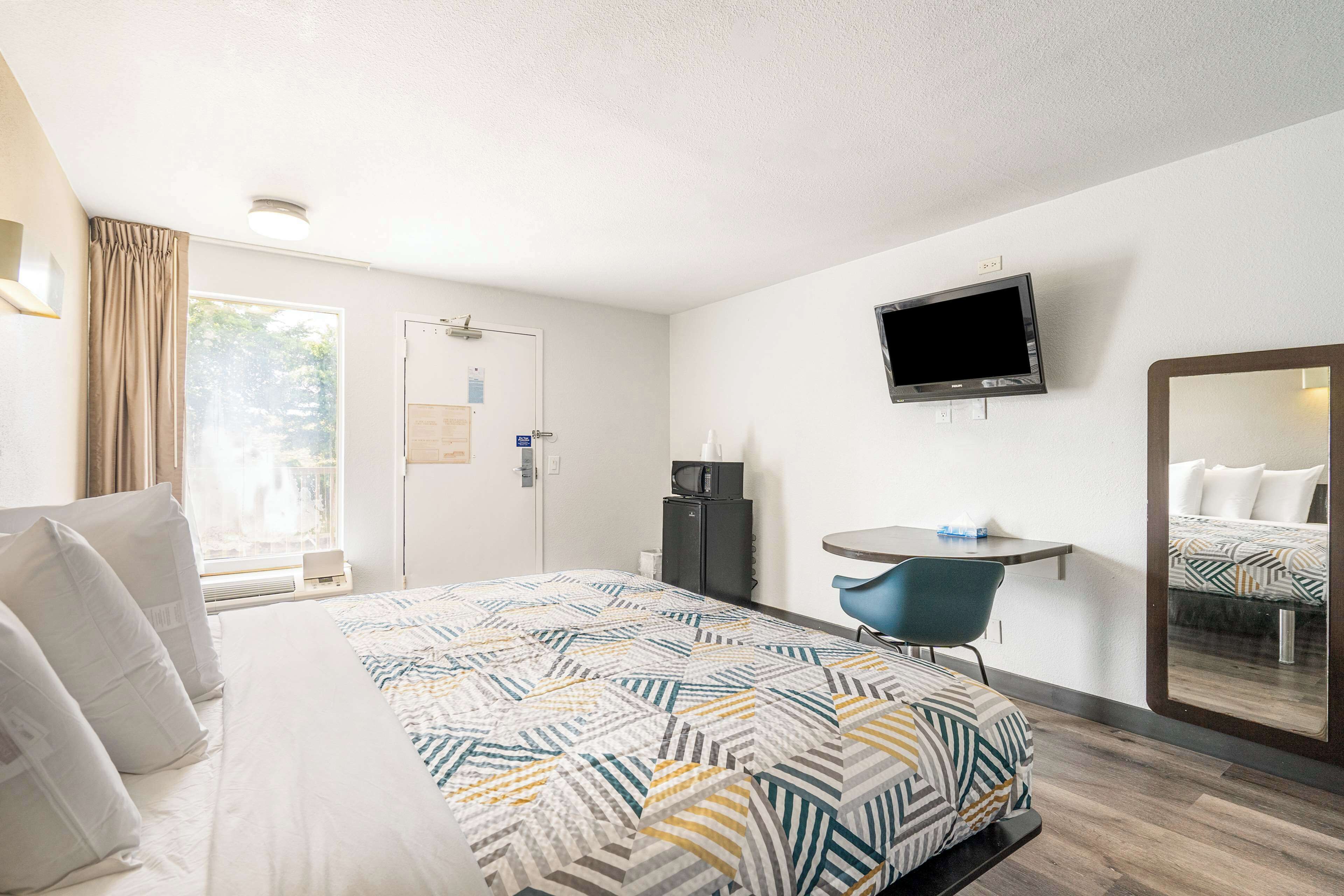 Habitación de hotel con cama, escritorio, silla, televisor y espejo.