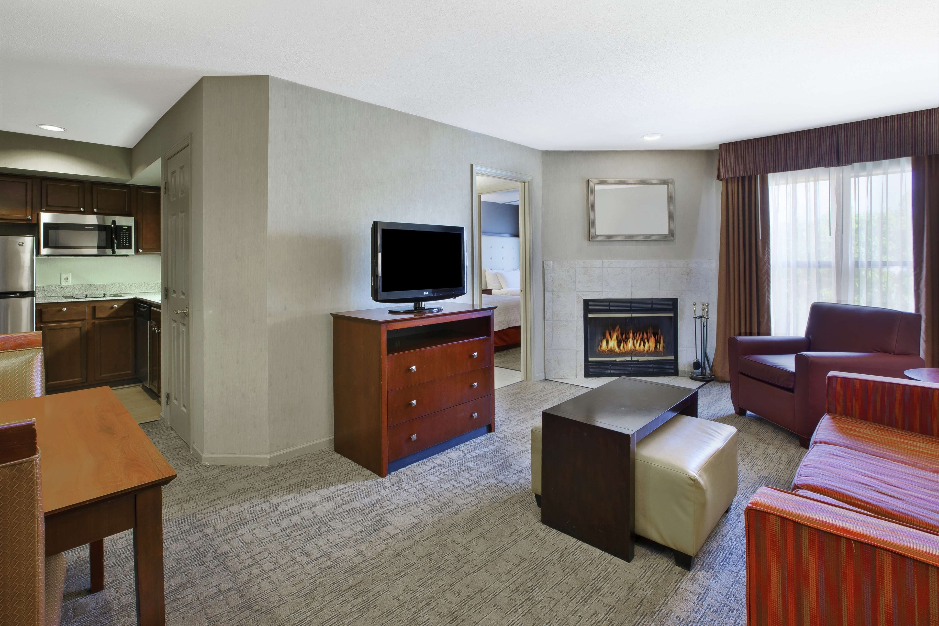 A hotel room with a kitchenette, a flat-screen TV, a fireplace, and a seating area with a sofa and armchairs.