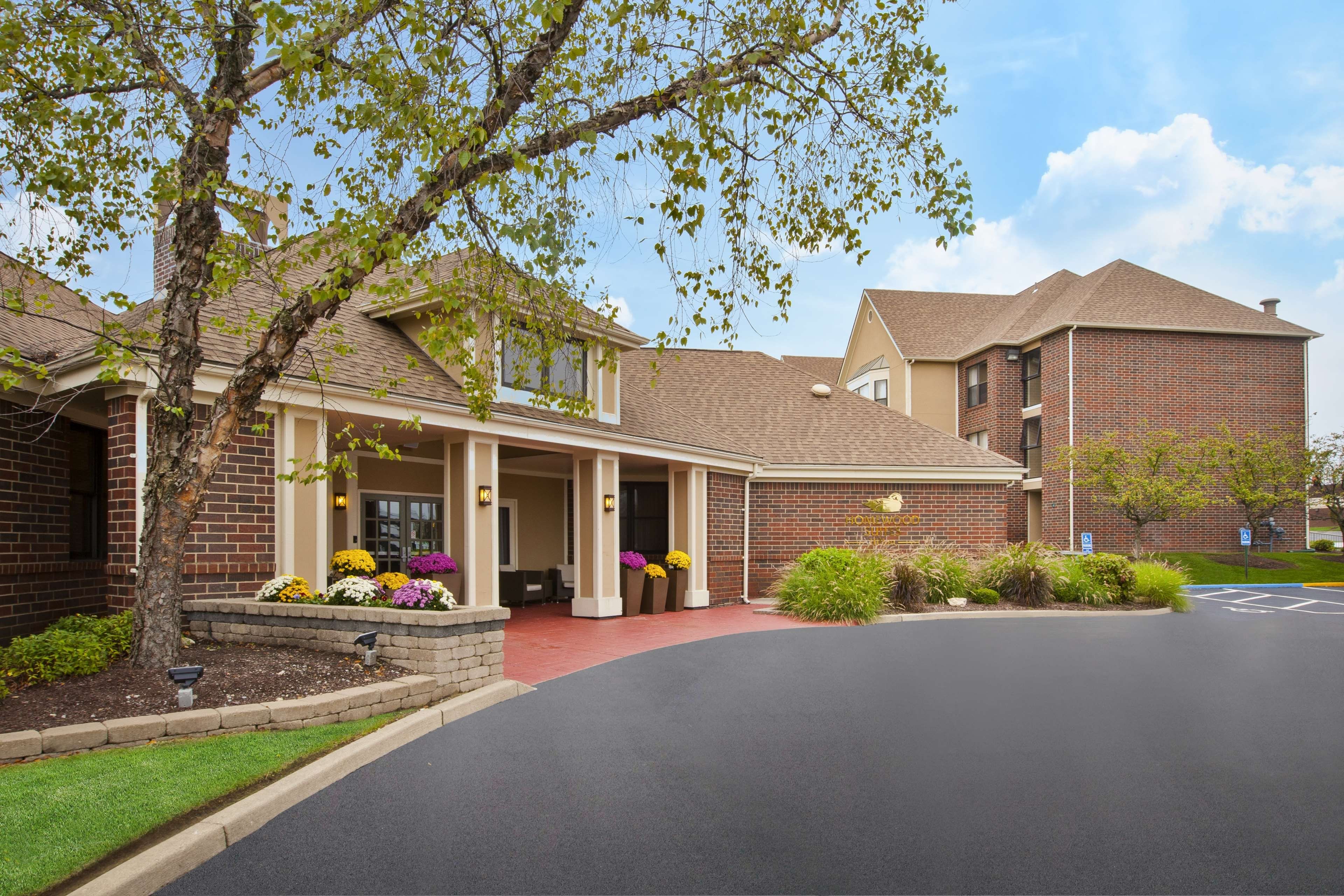 A two-story building with a brick facade, a covered entrance with a red carpet, and a landscaped front yard with various plants and flowers.