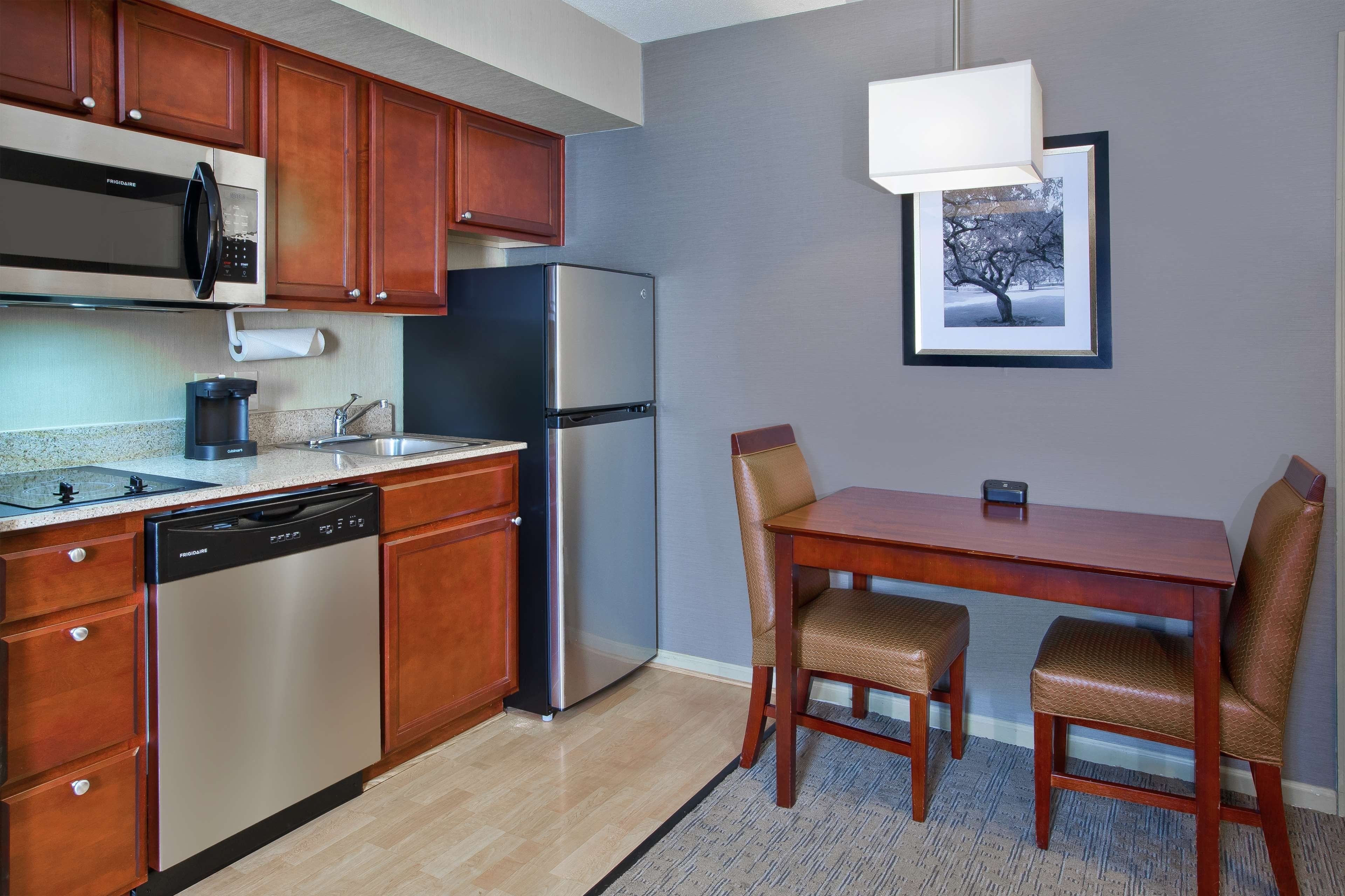 A compact kitchen with a dishwasher, a refrigerator, and a microwave. There is a dining table with two chairs and a framed picture on the wall.