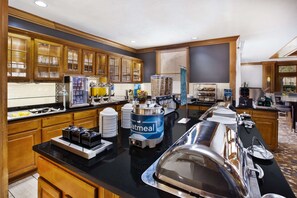 A hotel breakfast area with self-serve stations, including a coffee machine, a waffle station, and a fruit bowl.