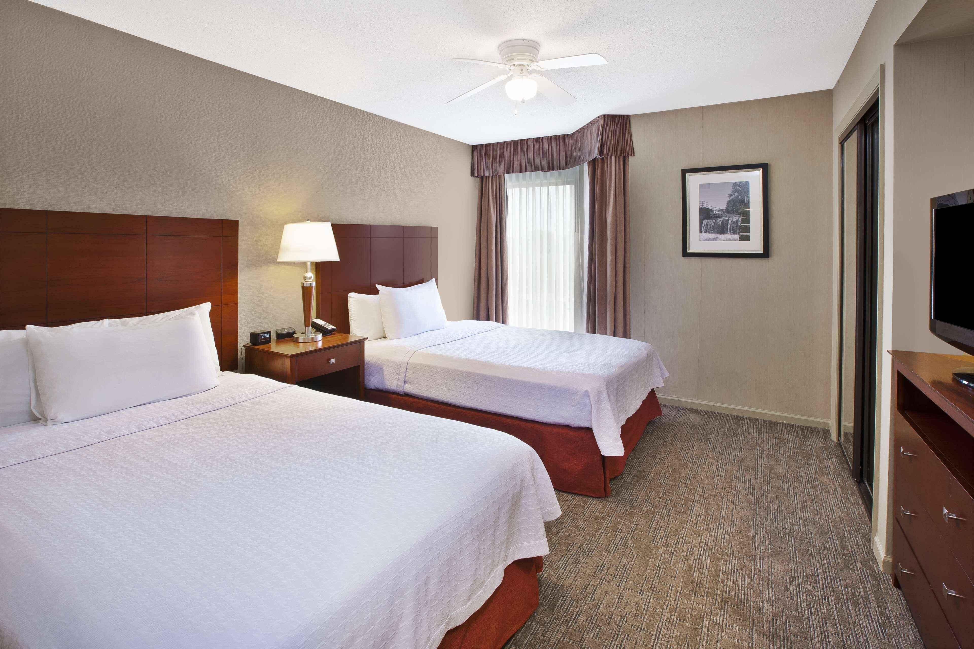 Une chambre d’hôtel avec deux lits, une table de chevet avec une lampe, un ventilateur de plafond, un tableau encadré au mur et un téléviseur posé sur une commode.