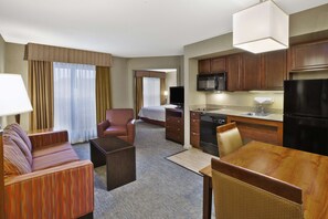 Quarto de hotel com sofá, poltrona, mesa de jantar, área de cozinha e uma cama.