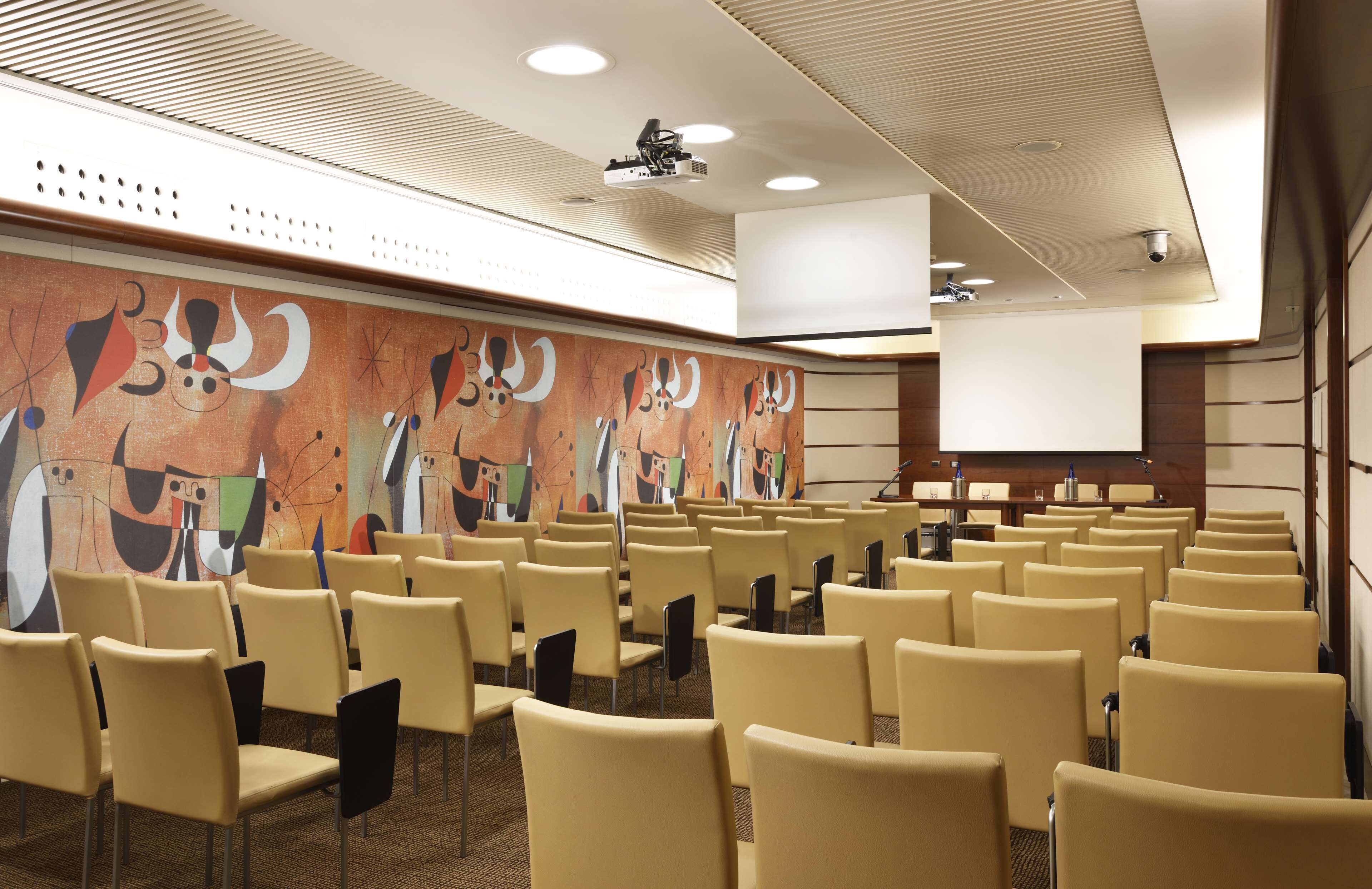 Ein Konferenzraum mit Reihen von Stühlen, einer großen Projektionsleinwand und abstrakter Wandkunst.