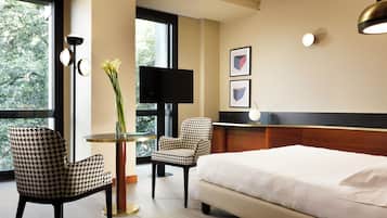 A hotel room with a bed, two houndstooth patterned chairs, a round table with a vase of flowers, a flat-screen TV, and framed artwork on the wall.