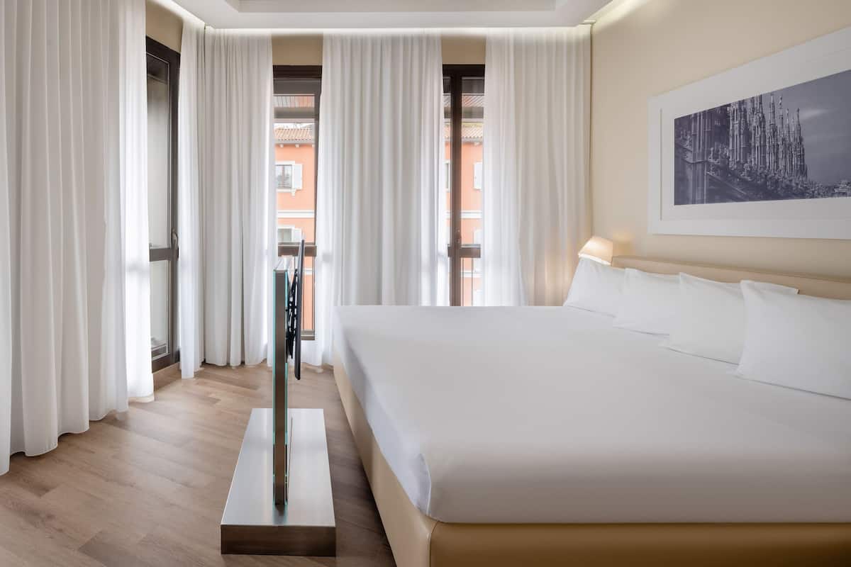 A hotel room with a large bed, white curtains, a framed picture on the wall, and a view of buildings outside.