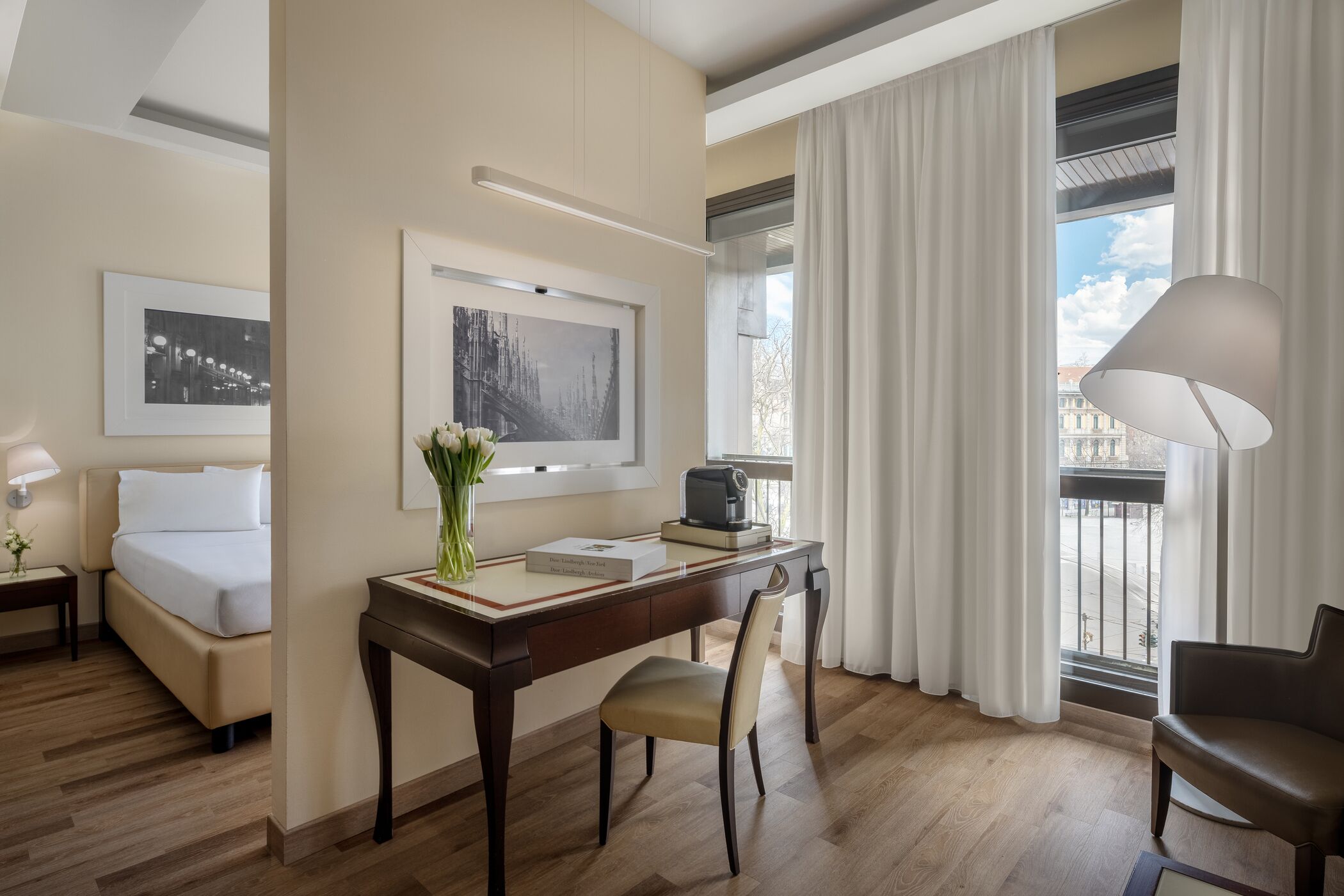 Une chambre d’hôtel équipée d’un lit, d’un bureau, d’une chaise et offrant une vue sur la ville grâce à de grandes fenêtres.