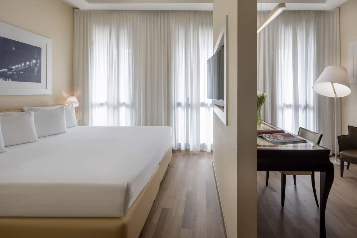 A hotel room with a large bed, a desk, and a chair. There is a TV mounted on the wall and a lamp on the desk.