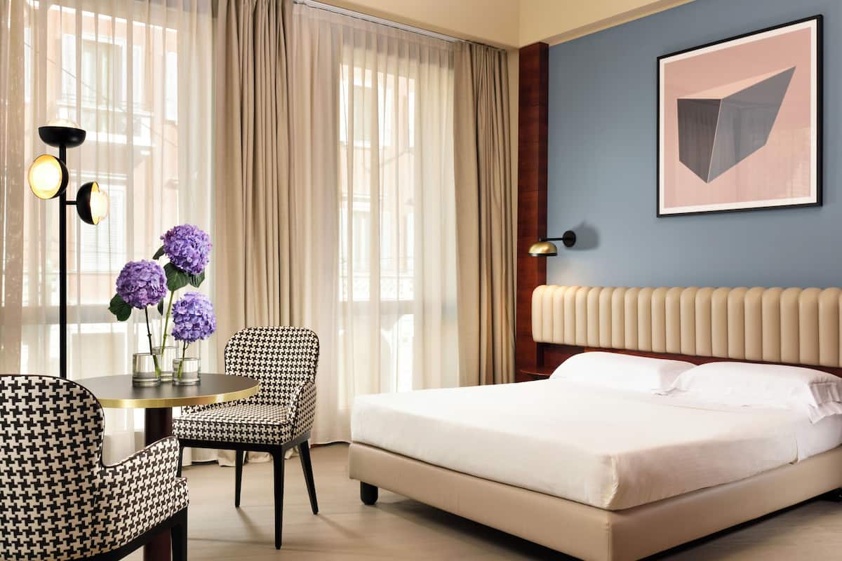 A hotel room with a bed, a chair, a table with a vase of flowers, and a lamp.