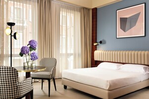 Een hotelkamer met een bed, een stoel, een tafel met een bloevas en een lamp.