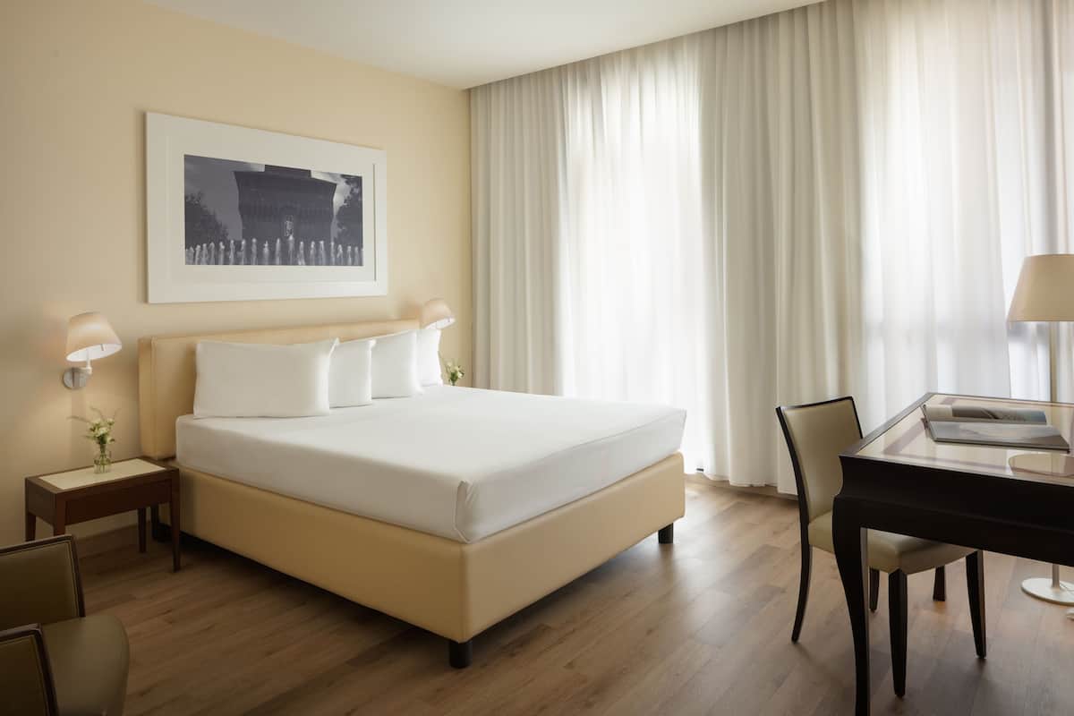 A hotel room with a large bed, a desk, and a chair. There is a framed picture on the wall and a small table with a lamp.
