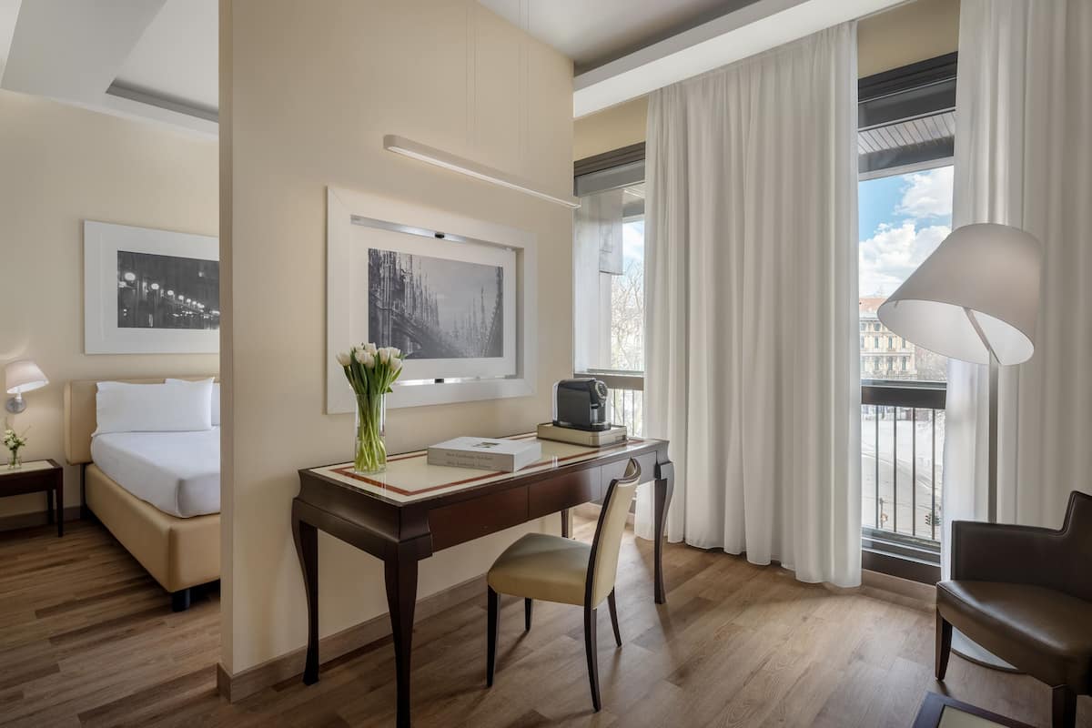 A hotel room with a bed, desk, chair, and a view of the city through large windows.