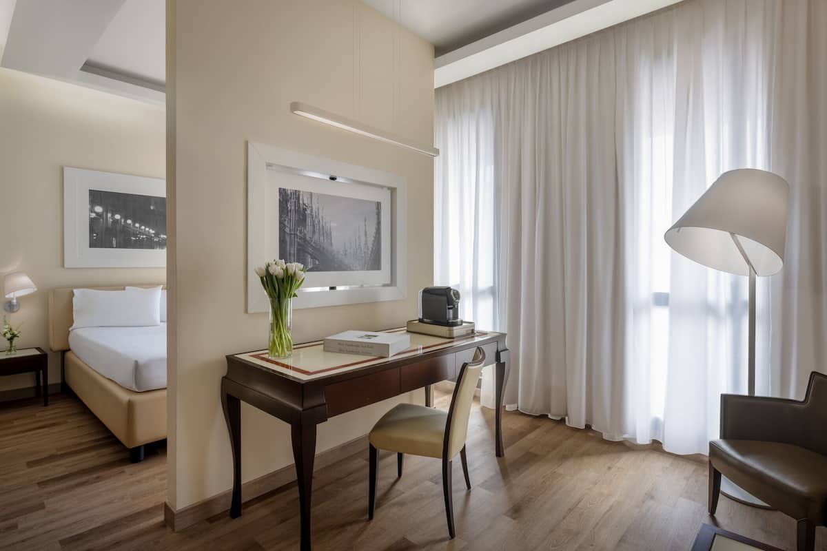 A hotel room with a bed, desk, chair, lamp, and artwork on the walls.