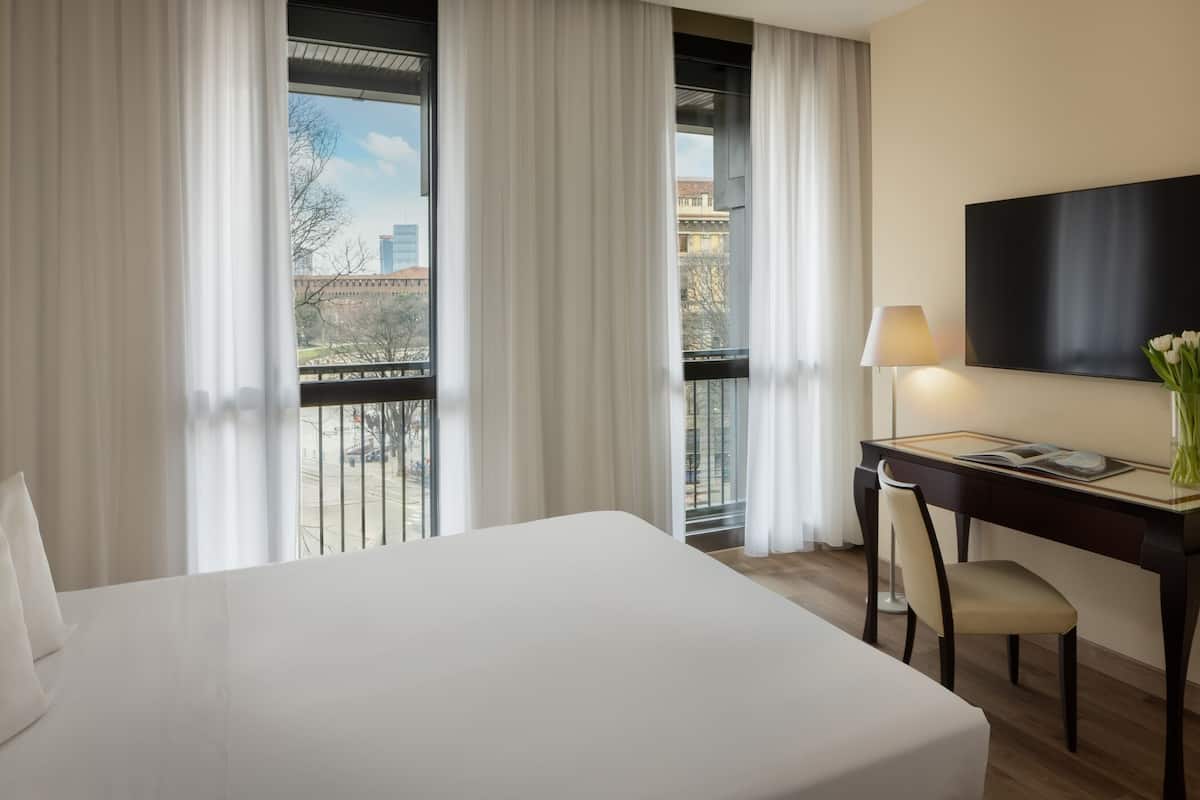 A hotel room with a bed, a desk with a chair, a TV, and a view of the city through large windows.