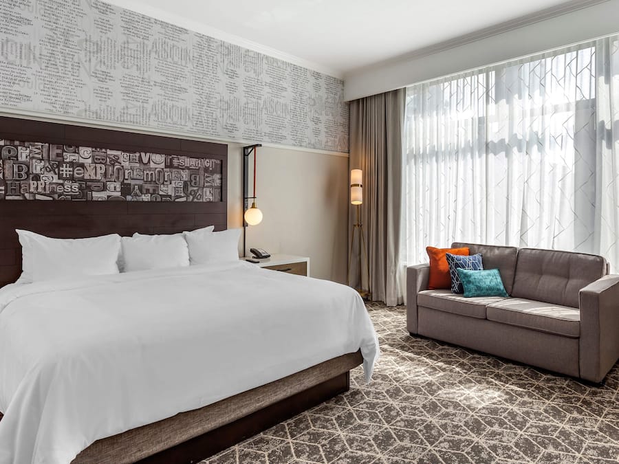 Habitación de hotel con una cama grande, un sofá, una mesita de noche, una lámpara y una ventana con cortinas.