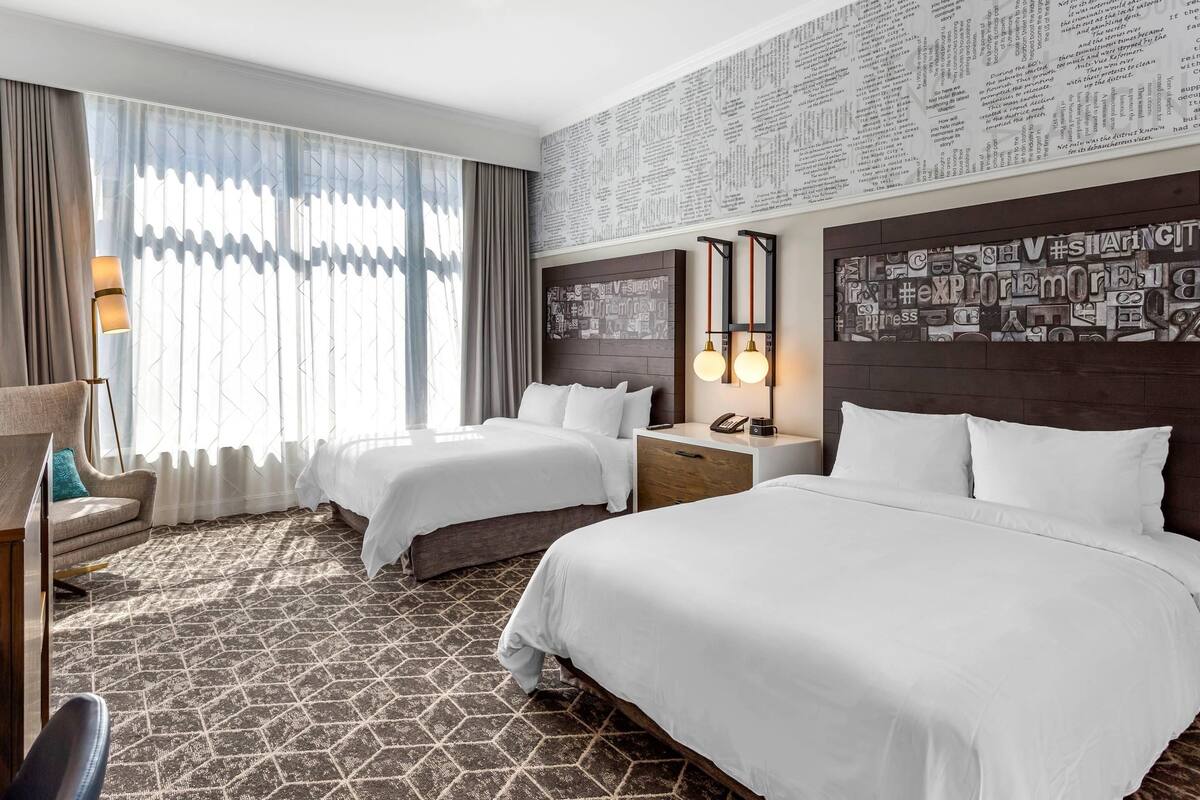 Habitación de hotel con dos camas, un escritorio, una silla y un ventanal grande con cortinas.