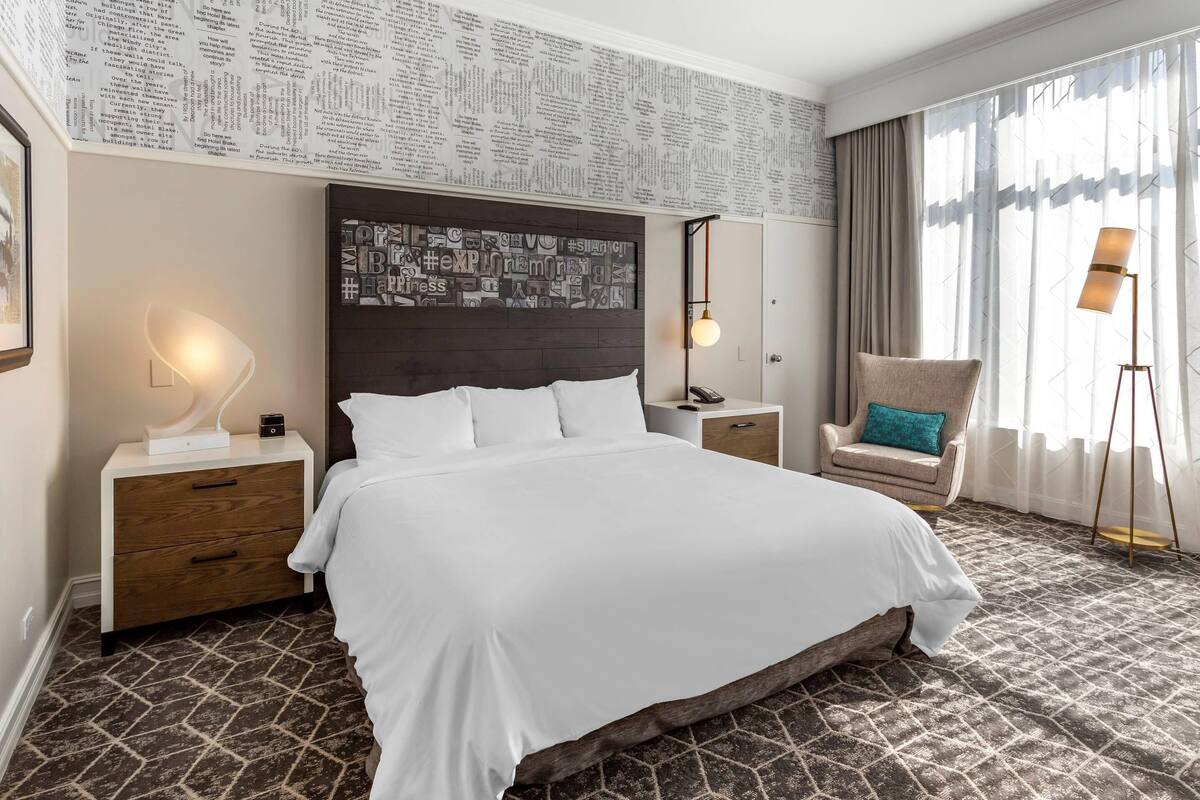 Una habitación de hotel con una cama grande, una mesita de noche de madera, una silla con un cojín color turquesa, una lámpara de pie y una pared con papel tapiz de periódico.