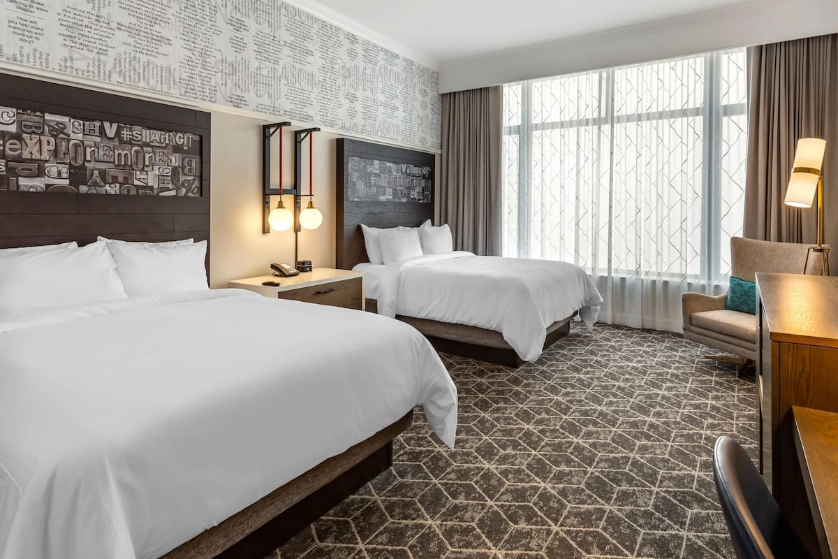 Habitación de hotel con dos camas, un ventanal con cortinas, una pared con recortes de periódico y un escritorio con lámpara.