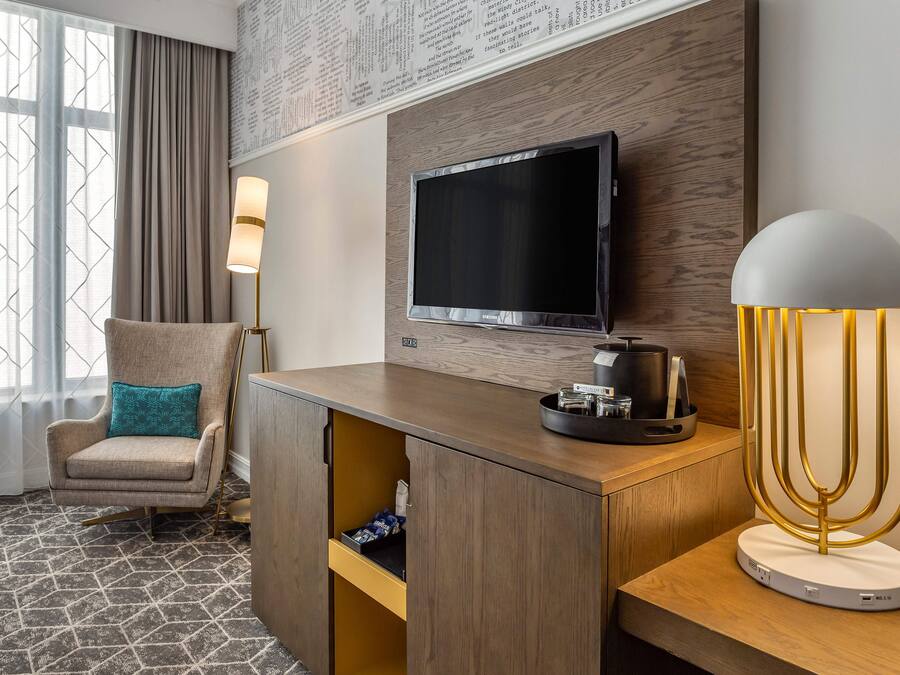 Une chambre d’hôtel dotée d’un téléviseur à écran plat fixé à un mur en bois, d’un canapé agrémenté d’un coussin turquoise, d’un bureau avec une lampe et d’une machine à café.