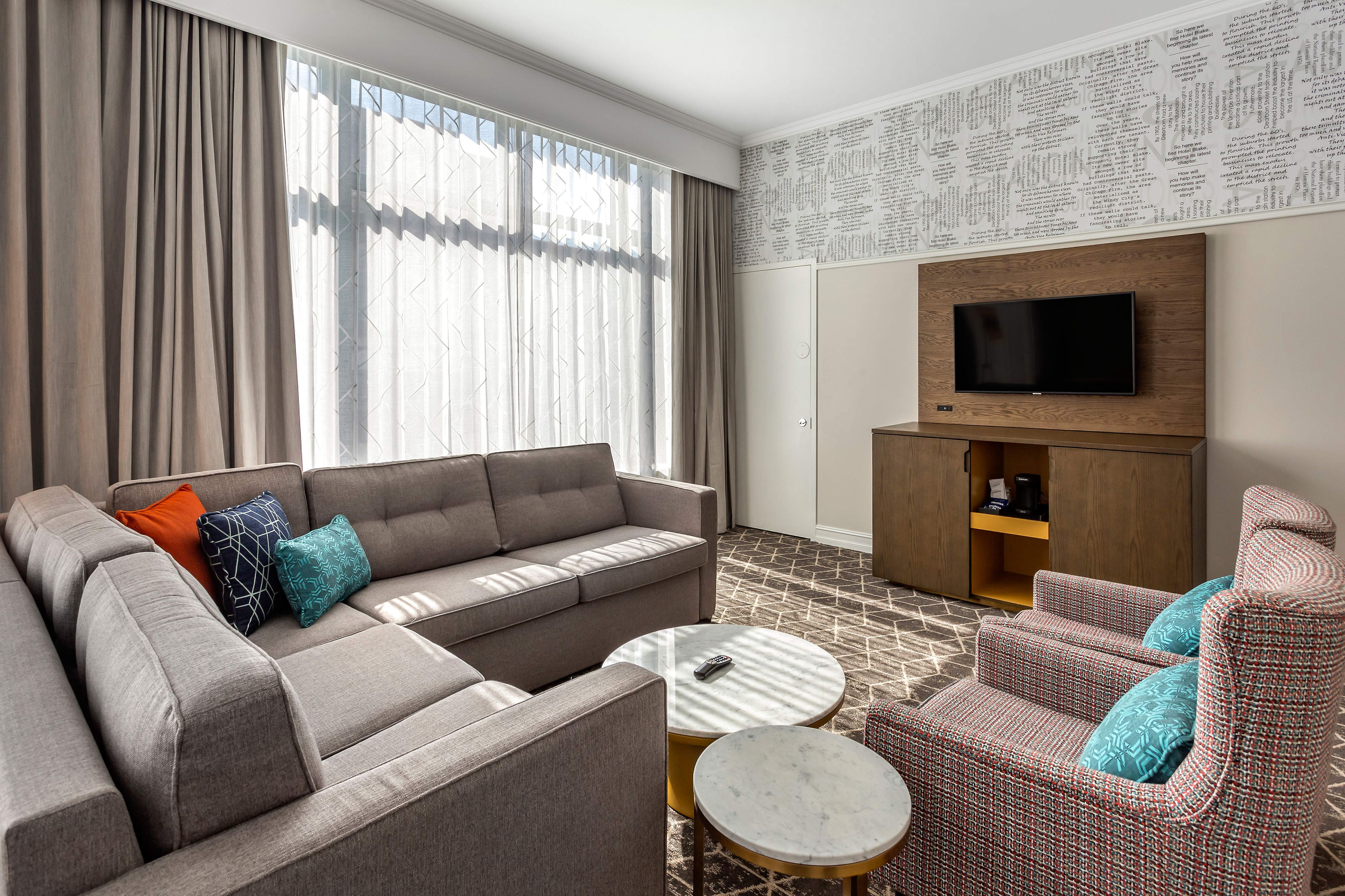 A modern hotel room with a sofa, armchairs, a coffee table, a flat-screen TV, and a patterned wall.