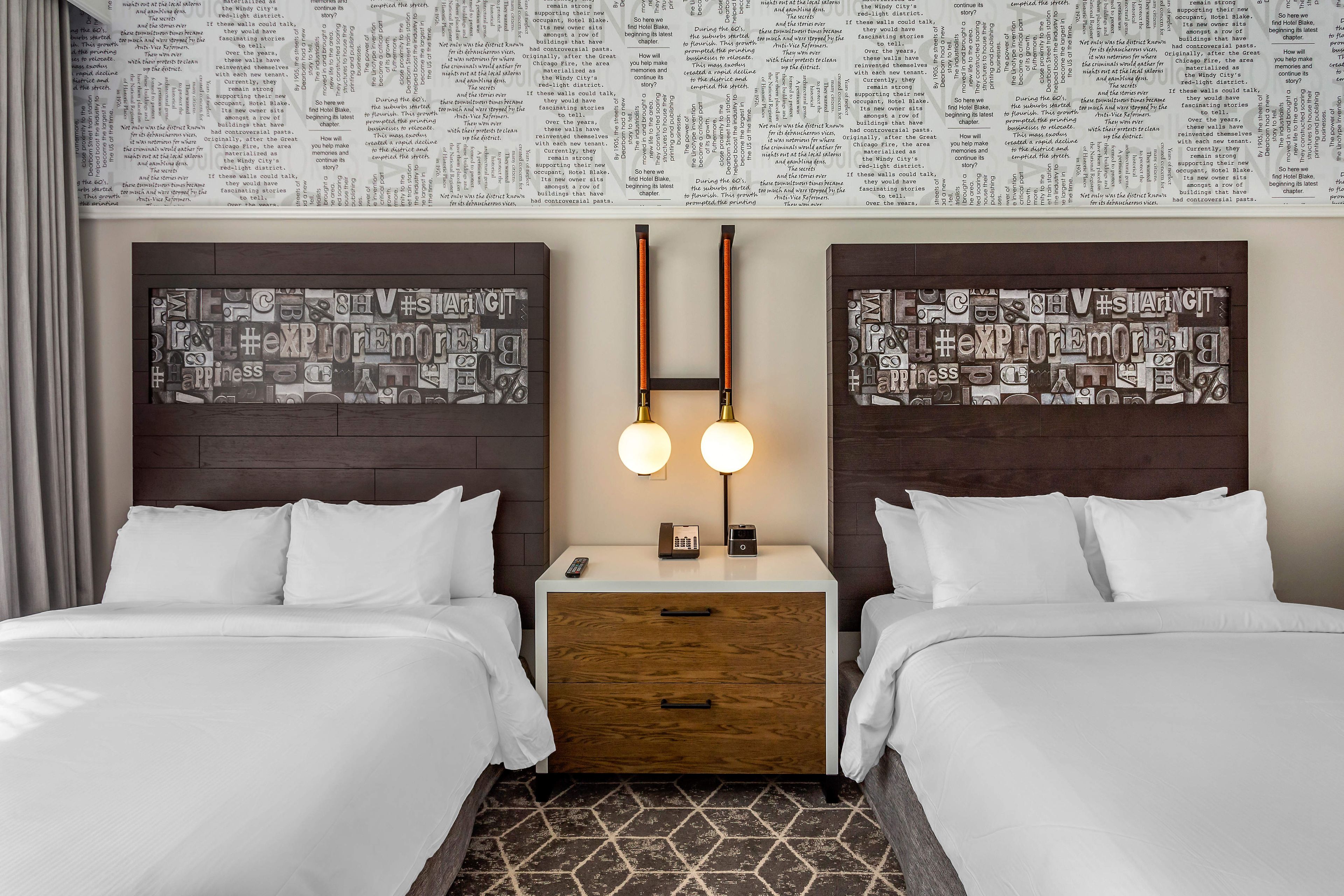 A hotel room with two beds, a nightstand with a lamp, and a wall with printed text.