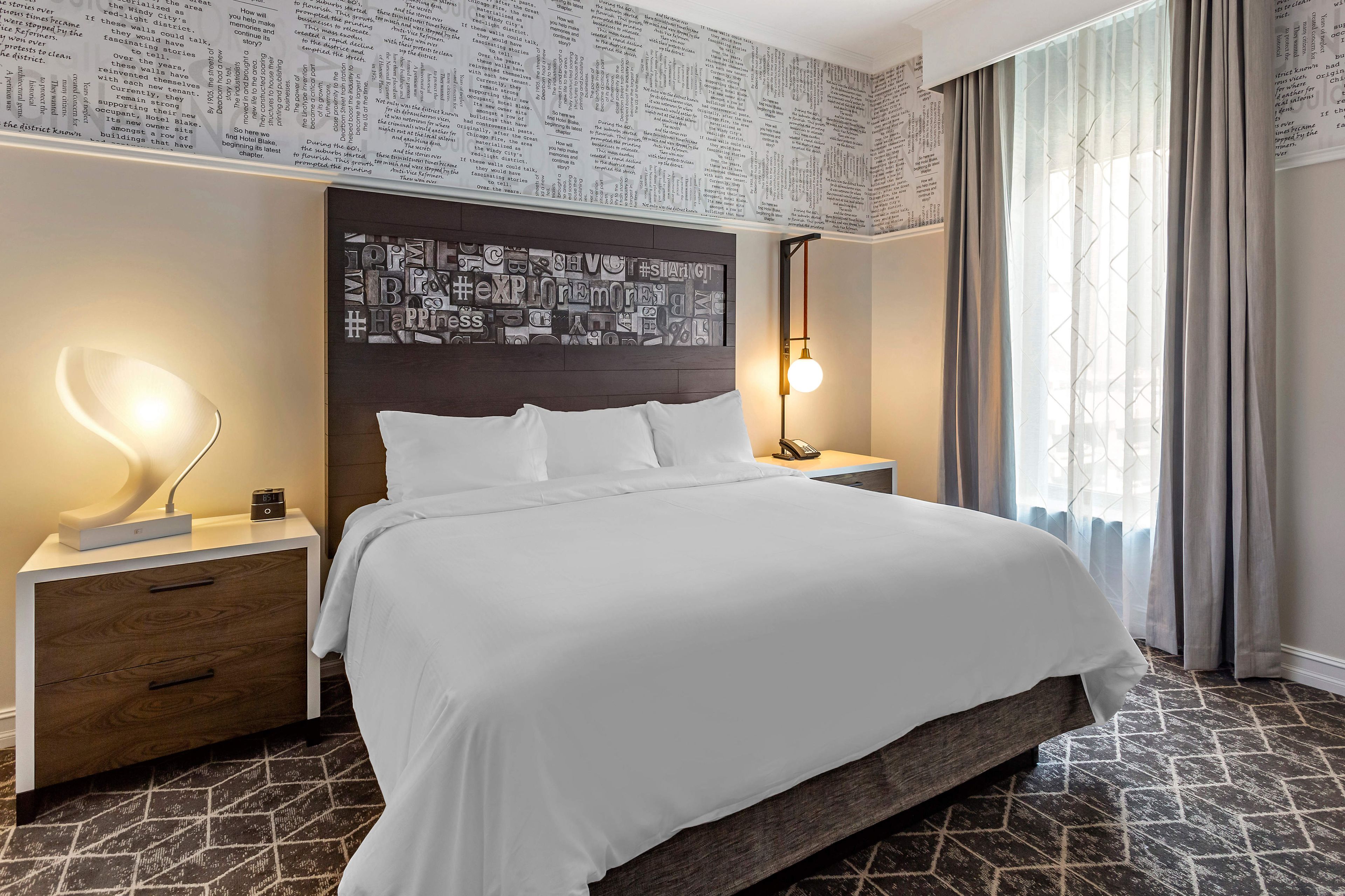 A hotel room with a large bed, a nightstand with a lamp, a wall clock, and a window with curtains.