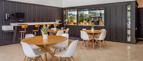 A modern dining area with round tables and white chairs, a kitchen area with a coffee machine, and a view of the outdoors through large windows.