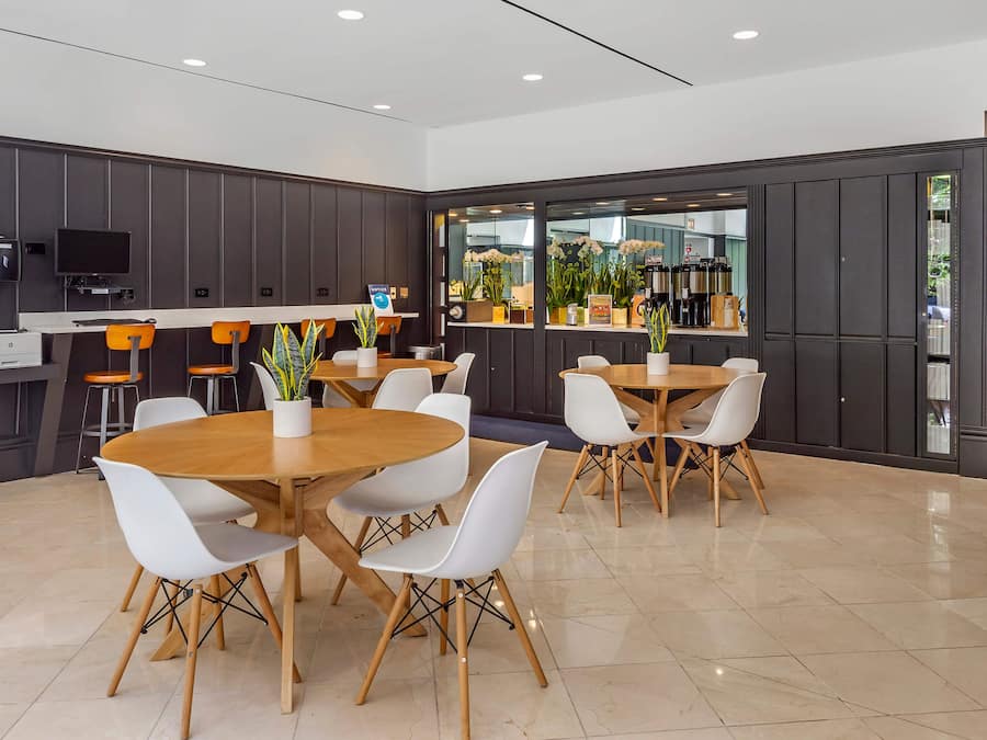 A modern dining area with round tables and white chairs, a kitchen area with a coffee machine, and a view of the outdoors through large windows.
