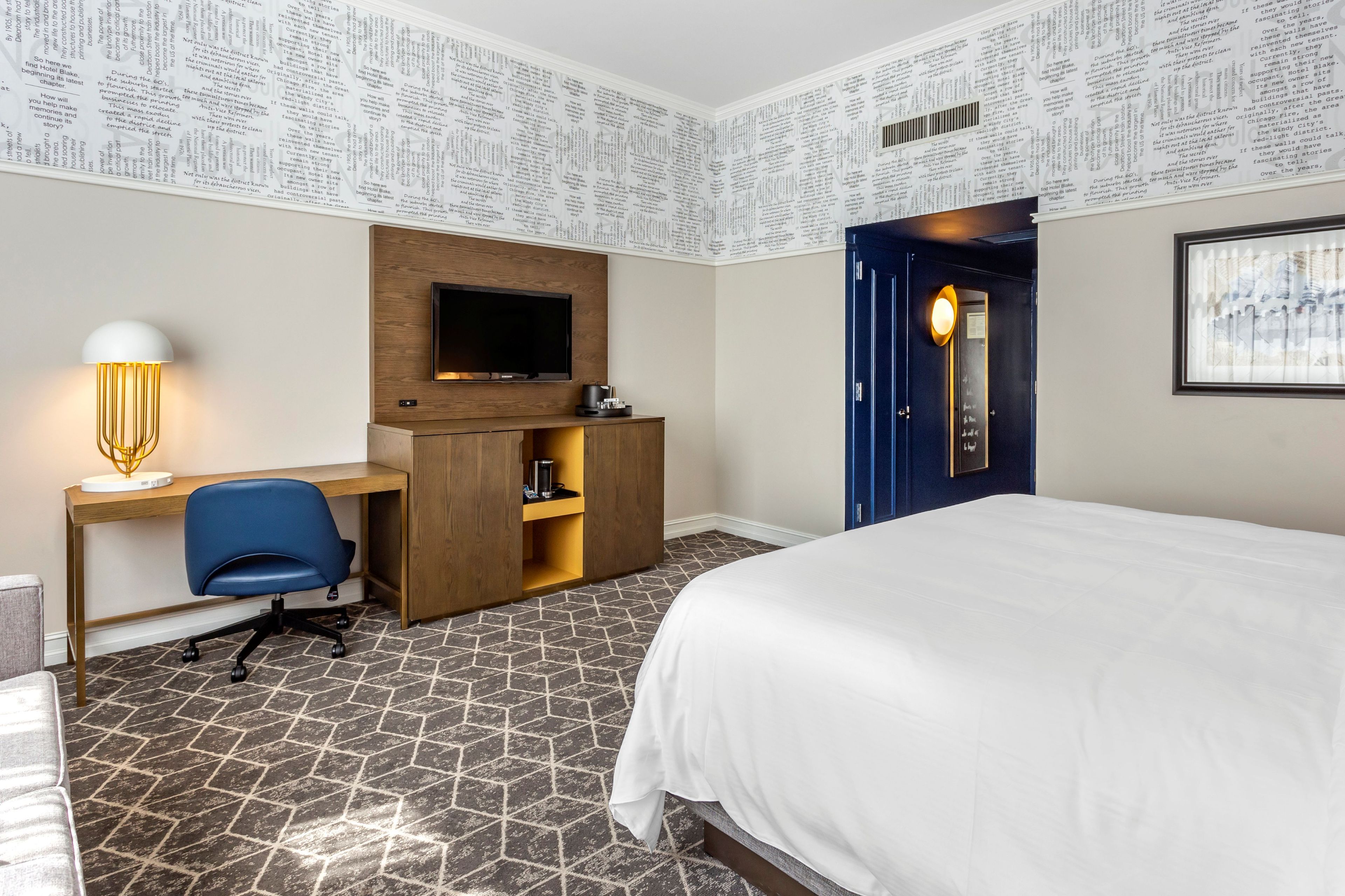 Una habitación de hotel con cama, escritorio con silla, televisor y puerta con luz.