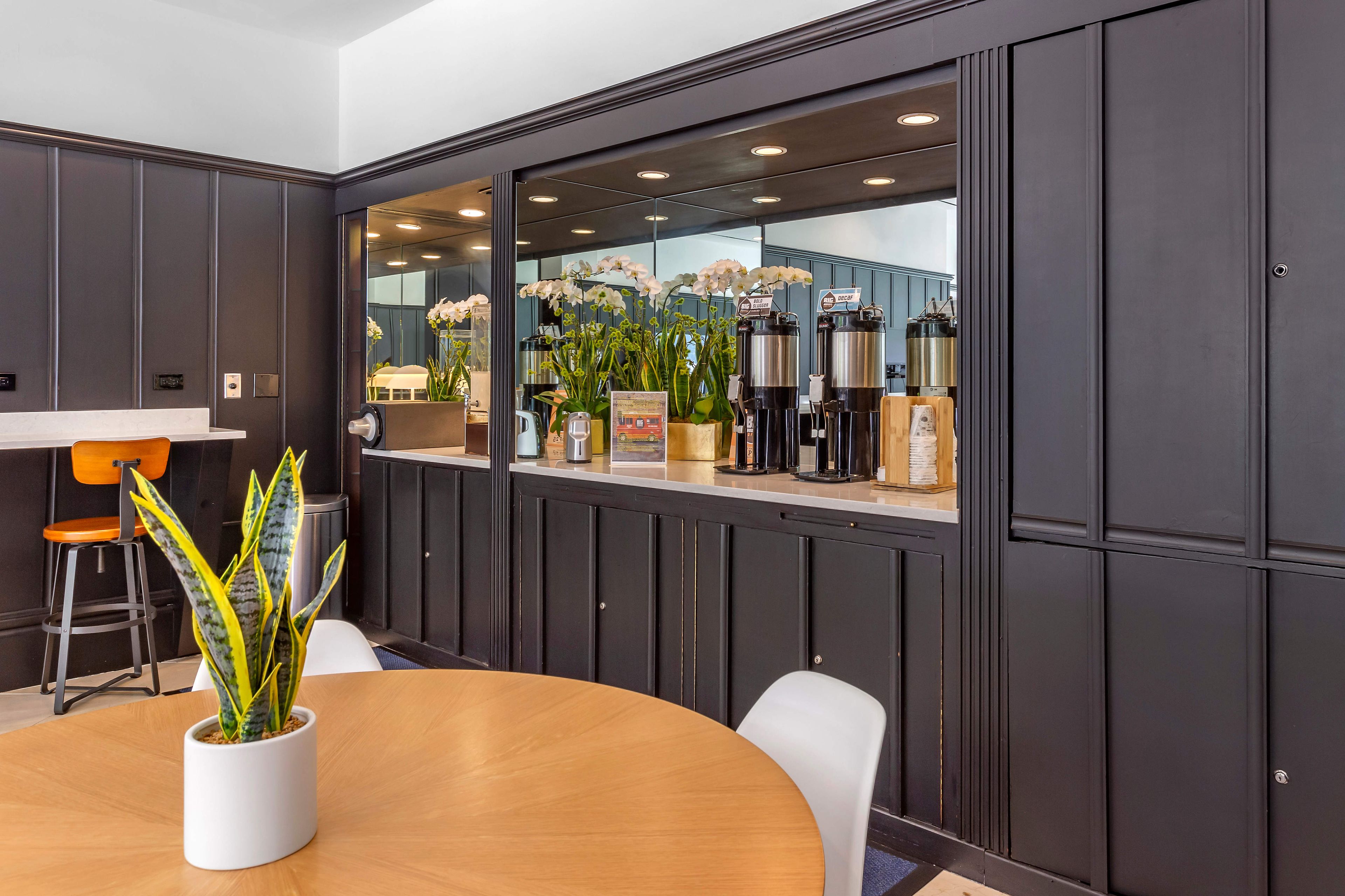 A modern conference room with a wooden table, white chairs, a potted plant, and a large window with decorative plants.
