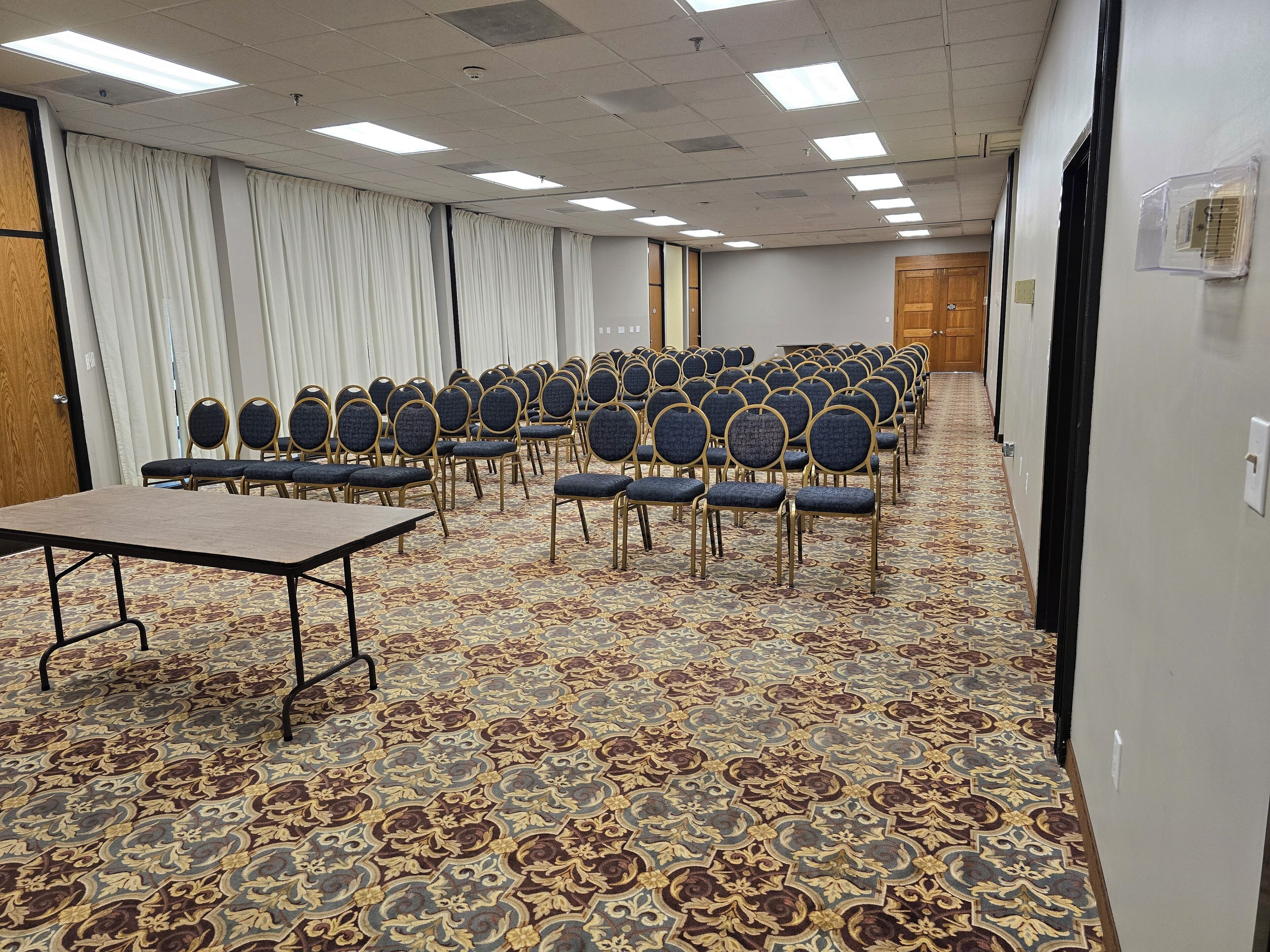 Un salón de conferencias con filas de sillas y una mesa, suelo alfombrado e iluminación en el techo.