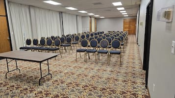 Un salón de conferencias con filas de sillas y una mesa, suelo alfombrado e iluminación en el techo.