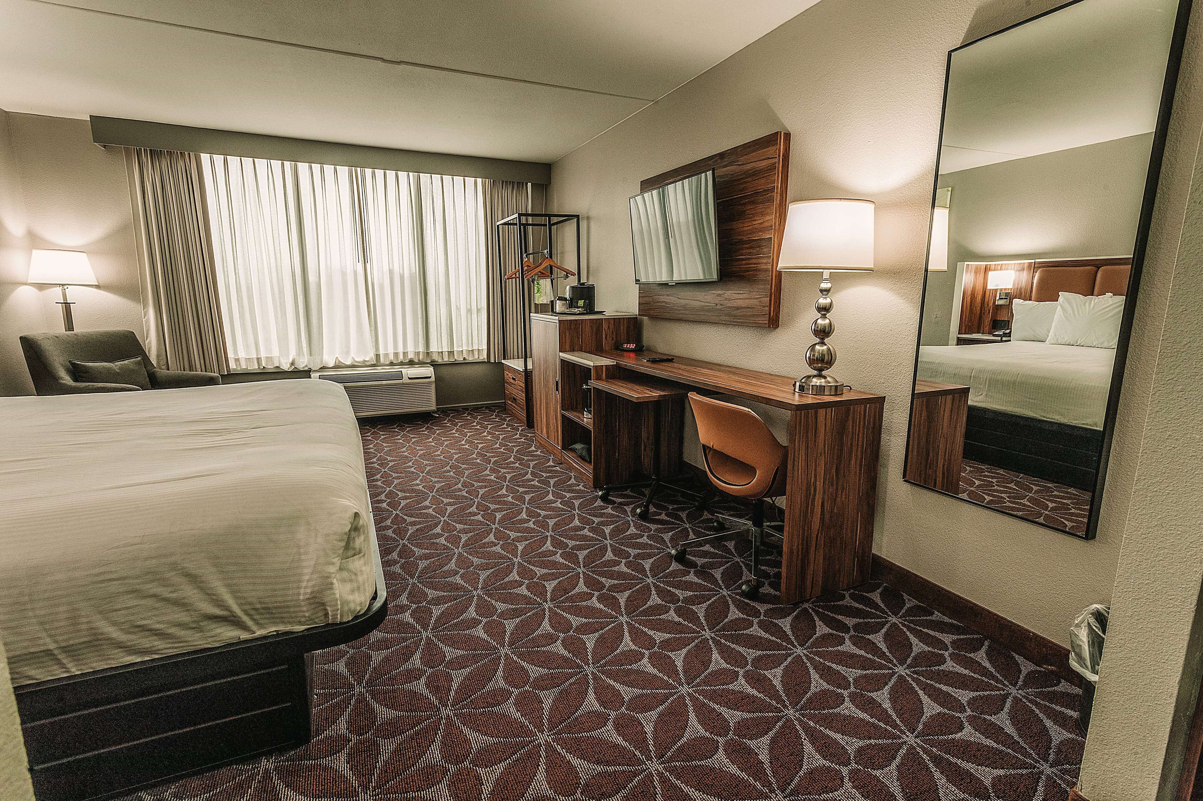 A hotel room with a large bed, a desk, a chair, a desk lamp, a mirror, and a window with curtains.