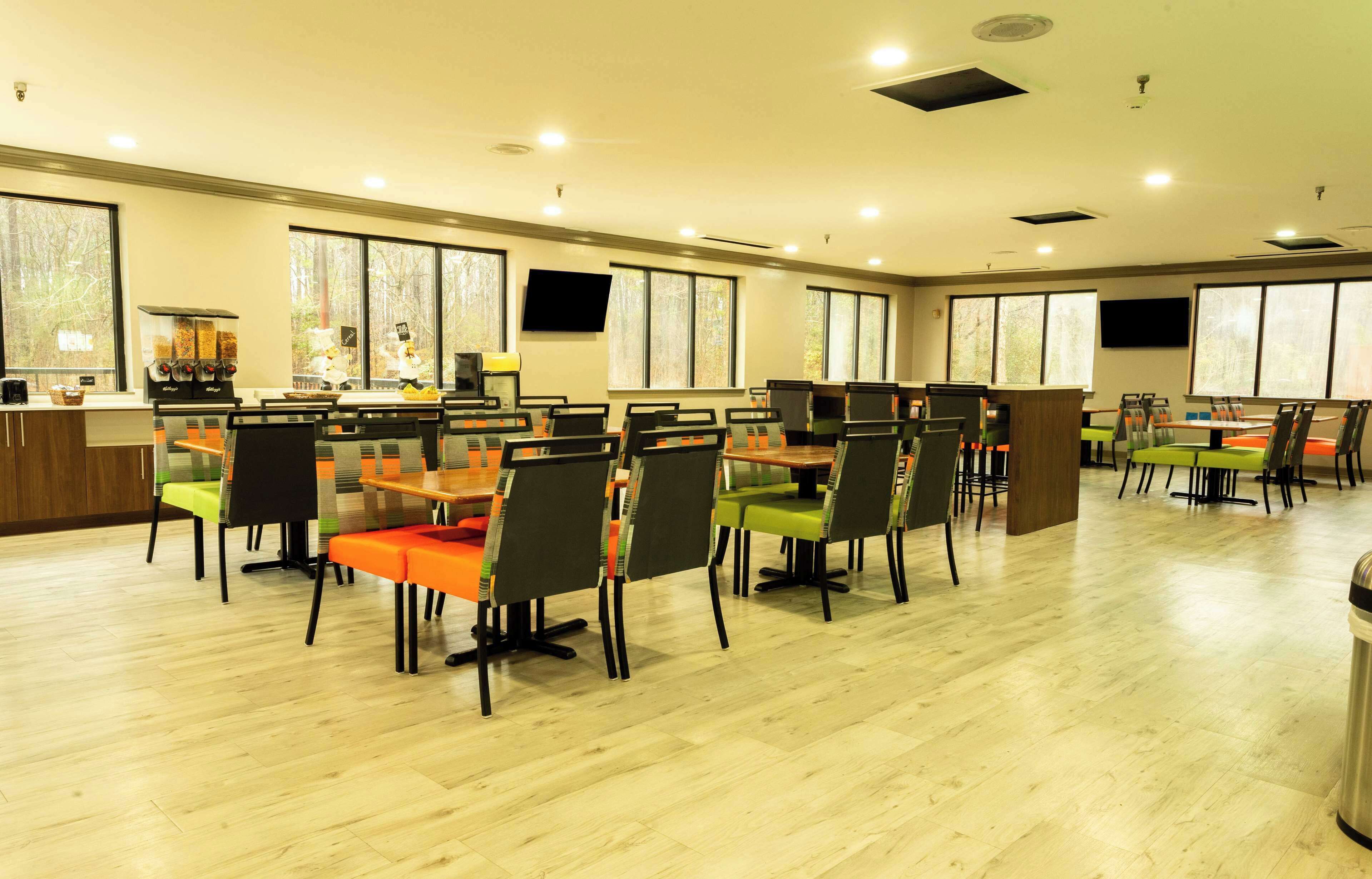 Uma ampla área de jantar com várias mesas e cadeiras, uma máquina de café e grandes janelas que proporcionam iluminação natural.