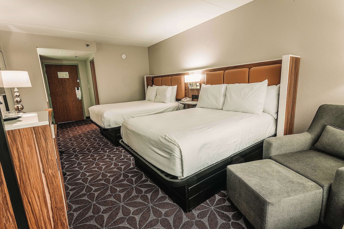 A hotel room with two beds, a desk, a chair, and a patterned carpet.