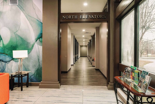 A hotel corridor with a marble floor, a table with a lamp, a bench with brochures, and a large window with a view.