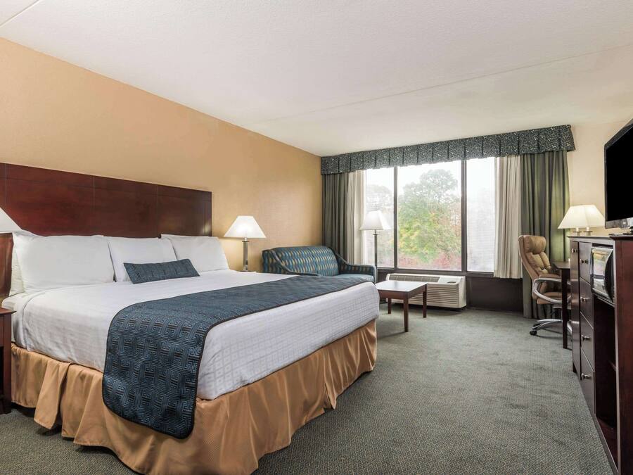 Ett hotellrum med en stor säng, två sängbord med lampor, ett skrivbord med en stol, en tv och ett fönster med gardiner.