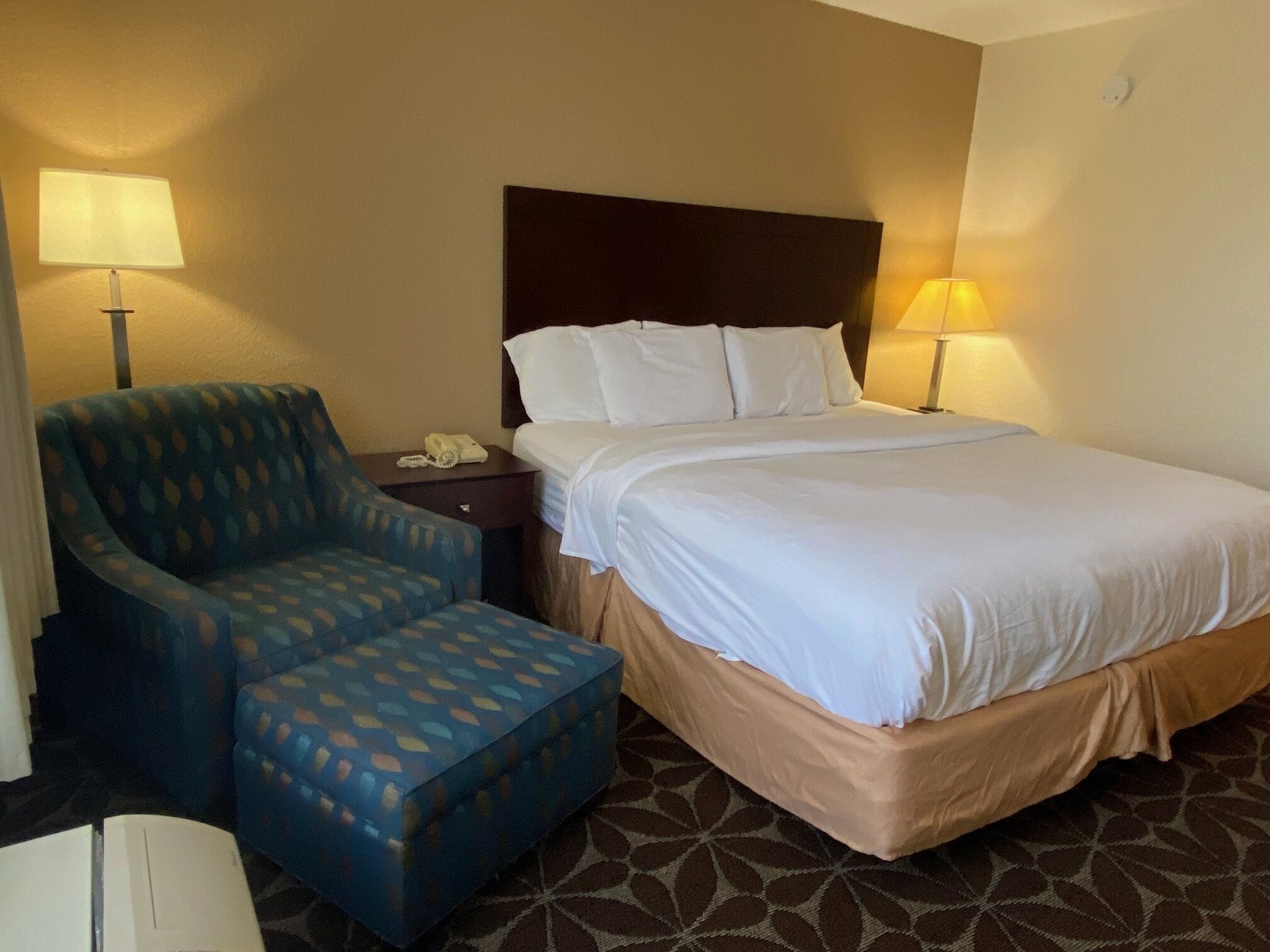Una habitación de hotel con una cama, una silla, una mesita de noche y dos lámparas.
