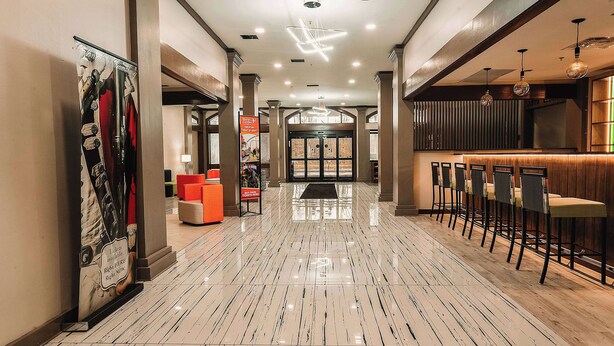 A hotel lobby with a polished marble floor, a reception area with a bar, and a large poster on the left wall.