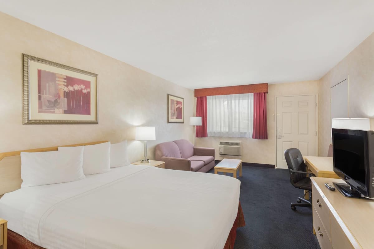 A hotel room with a bed, a desk with a computer, a TV, a chair, and a window with curtains.