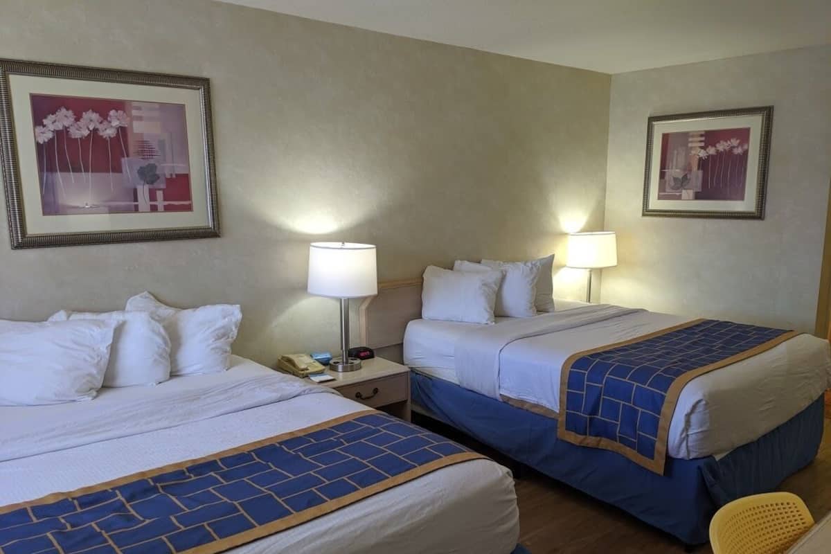 A hotel room with two beds, each with a blue and white bedspread, a bedside lamp, a nightstand with a phone, and a framed picture on the wall.