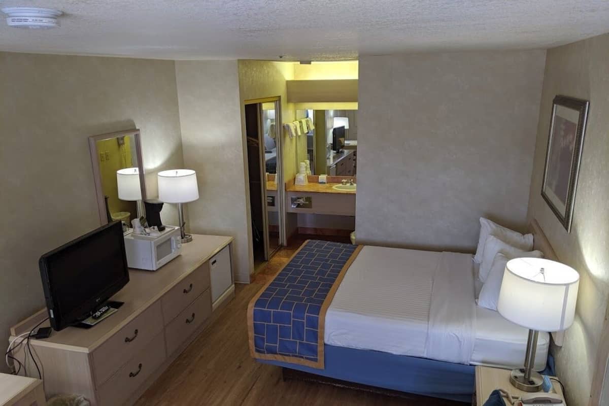 A hotel room with a bed, a television, a desk with drawers, a trash bin, and a bathroom visible through an open door.