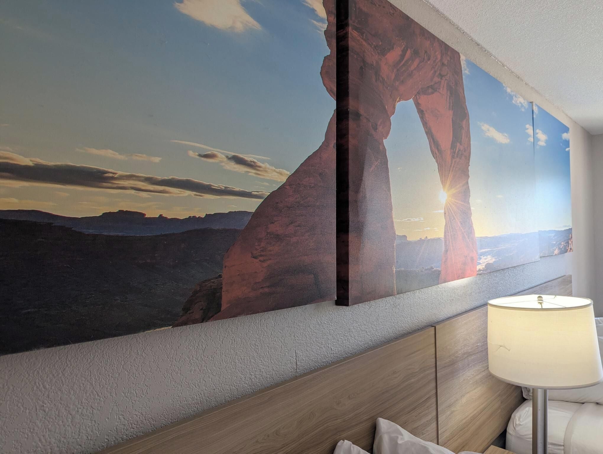 A hotel room with a large wall mural of a desert landscape, a wooden headboard, and a bedside lamp.
