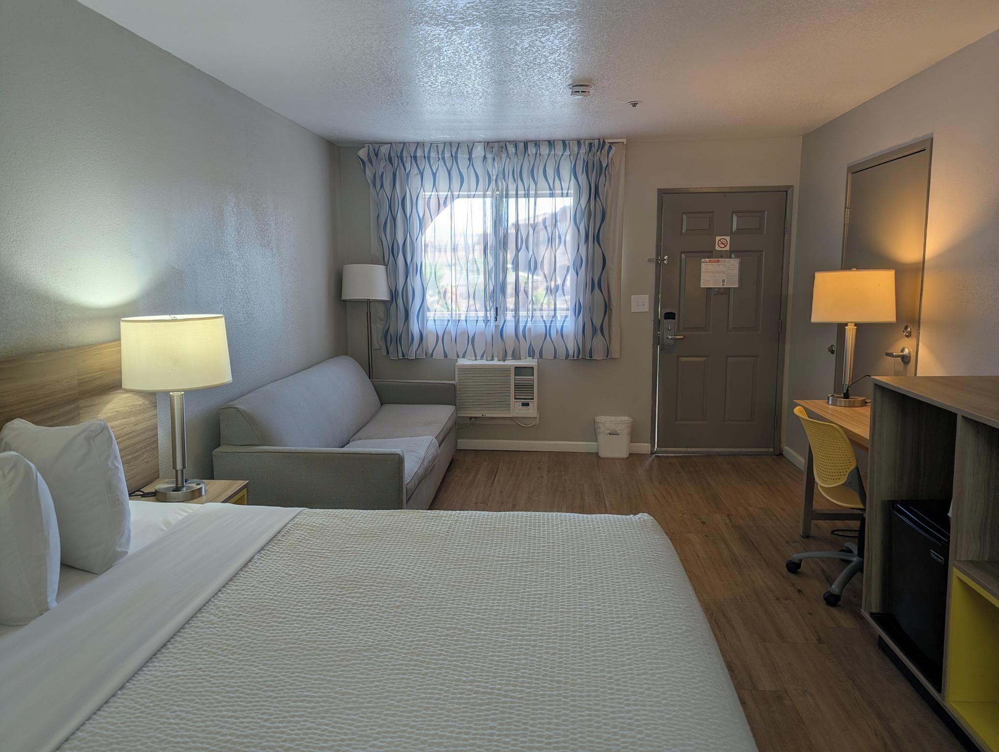 Et hotelværelse med seng, sofa, skrivebord med lampe, et vindue med gardiner og en dør.