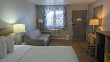 Et hotelværelse med seng, sofa, skrivebord med lampe, et vindue med gardiner og en dør.