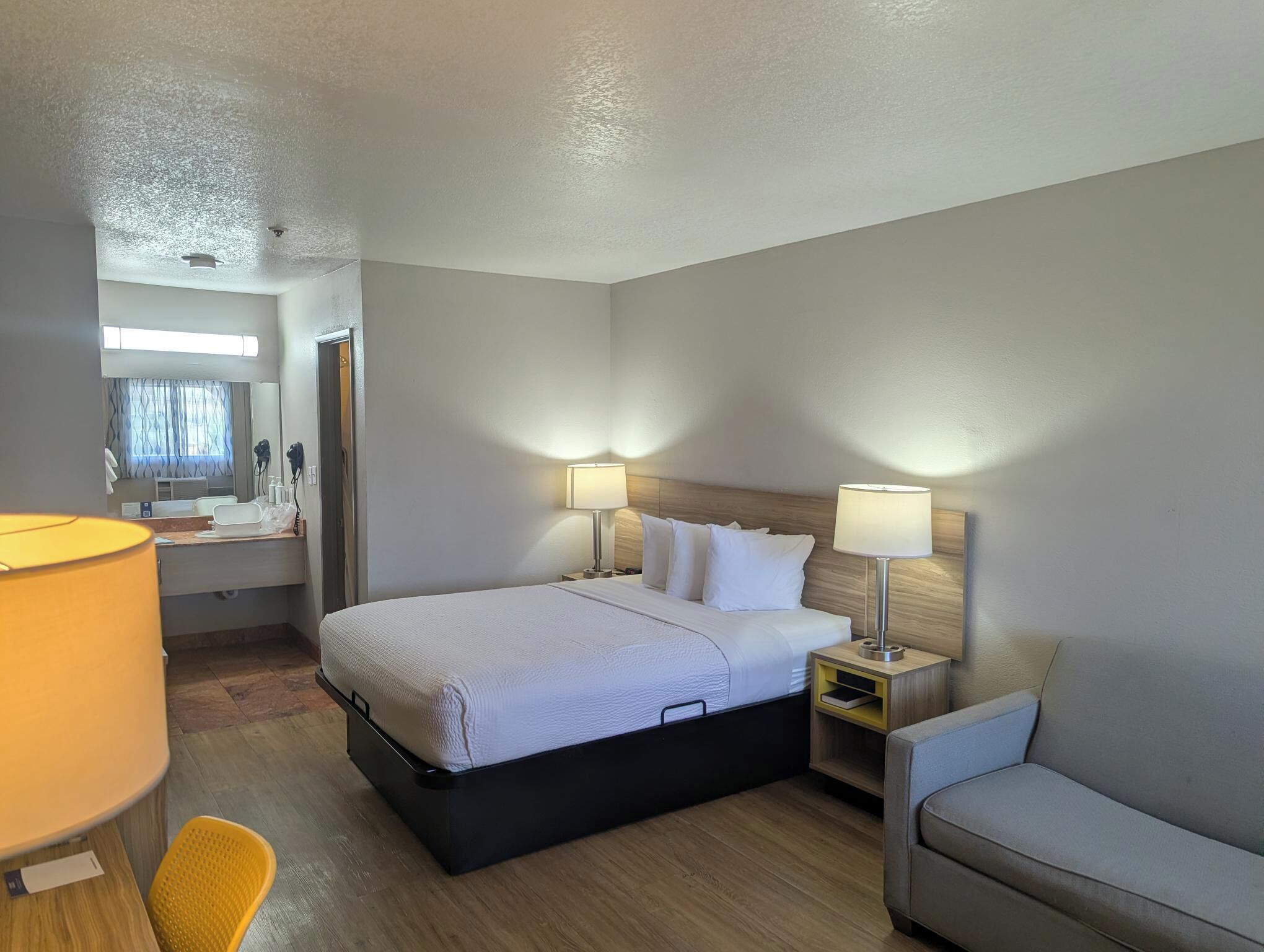 A hotel room with a bed, bedside lamps, a chair, a desk with a lamp, and a mirror.
