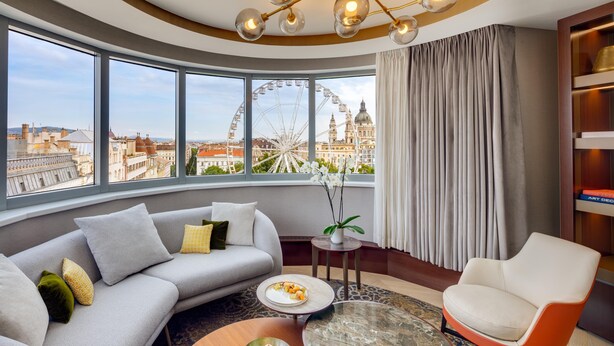 A modern living room with a curved sofa, a round coffee table, and a chair. A large window offers a view of a Ferris wheel and cityscape. - Kempinski Hotel Corvinus Budapest (Budapest)